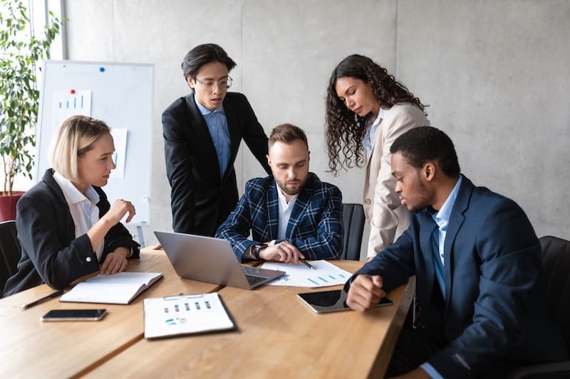 A diverse group of five professionals in business attire is gathered around a table in an office. Engaged in discussion, possibly on PR crisis management, they have charts and a laptop at hand. A whiteboard with graphs stands prominently in the background, supporting their analysis.