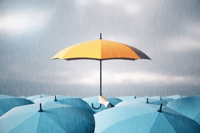 A cluster of blue umbrellas is shown in the rain. Among them, a single yellow umbrella, much like a pr crisis management strategy standing strong, stands out prominently in a person's hand, symbolizing individuality and contrast against the gray, rainy sky.