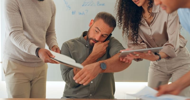 A man sits at a table looking tired and overwhelmed, rubbing his eyes. Three colleagues stand around him, armed with tablets and documents, entrenched in a PR crisis management meeting. A whiteboard with urgent notes looms in the background.