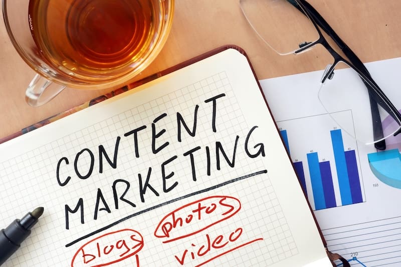 A notebook with "Content Marketing" scrawled across a page is surrounded by the words "blogs," "photos," and "video." Nearby, a pen, glasses, a cup of tea, and a chart with blue bars hint at branding in digital marketing strategies.