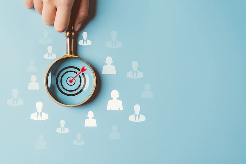 A hand holds a magnifying glass over a target with a red arrow at its center, emphasizing precision. White silhouettes of people fill the background, underscoring the concept of targeting in recruitment or branding in digital marketing.