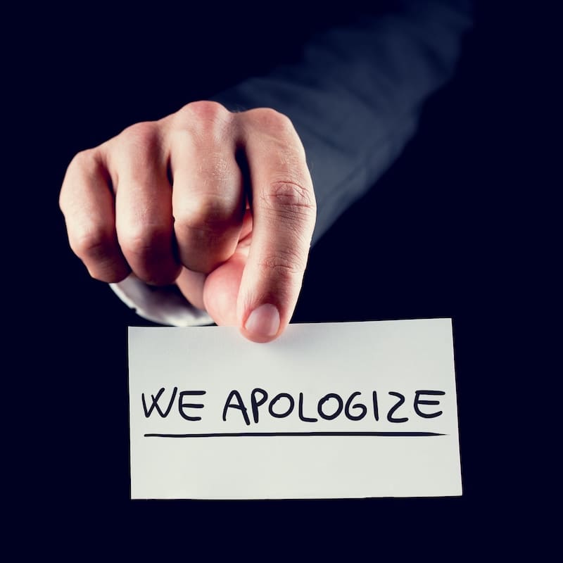 Close-up of a hand holding a white card with the words "We Apologize" in bold, black letters, reminiscent of an online message. The dark background highlights the card and its earnest apology.