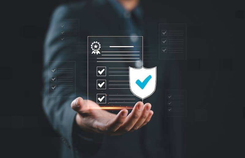 A person in a suit holds an online holographic checklist with a shield icon displaying a checkmark, symbolizing security and completion. The dark background accentuates the glowing effects of the hologram.