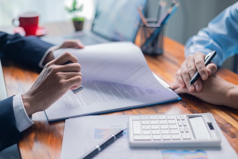 Two people in formal attire sit at a wooden table, reviewing documents. One, possibly an online reputation manager, holds a pen over a page while a calculator and charts rest nearby. A laptop and a red coffee cup are visible in the background, suggesting a business meeting.