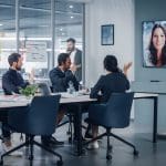 A group of five people in a modern conference room engages in a video call with an online reputation manager. Two individuals appear on a large screen. The room is well-lit and organized, featuring sticky notes on the glass wall and various office supplies on the tables.