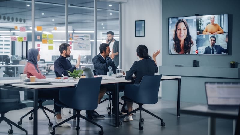 A group of five people in a modern conference room engages in a video call with an online reputation manager. Two individuals appear on a large screen. The room is well-lit and organized, featuring sticky notes on the glass wall and various office supplies on the tables.