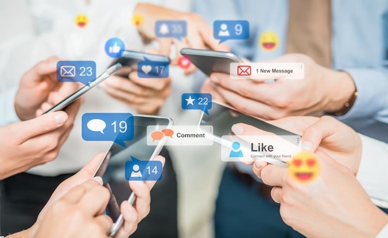 A group of people holding smartphones with a variety of social media notifications popping up around them, including likes, comments, messages, and emojis. Various hands are interacting with the devices, highlighting engagement in social media activities crucial for corporate reputation management.
