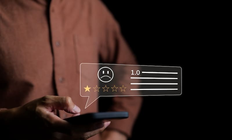 A person in a brown shirt holds a smartphone, with a transparent graphic overlay showing a 1-star rating out of 5, a sad face emoji, and a score of 1.0. The image highlights the critical need for corporate reputation management. The background is black.