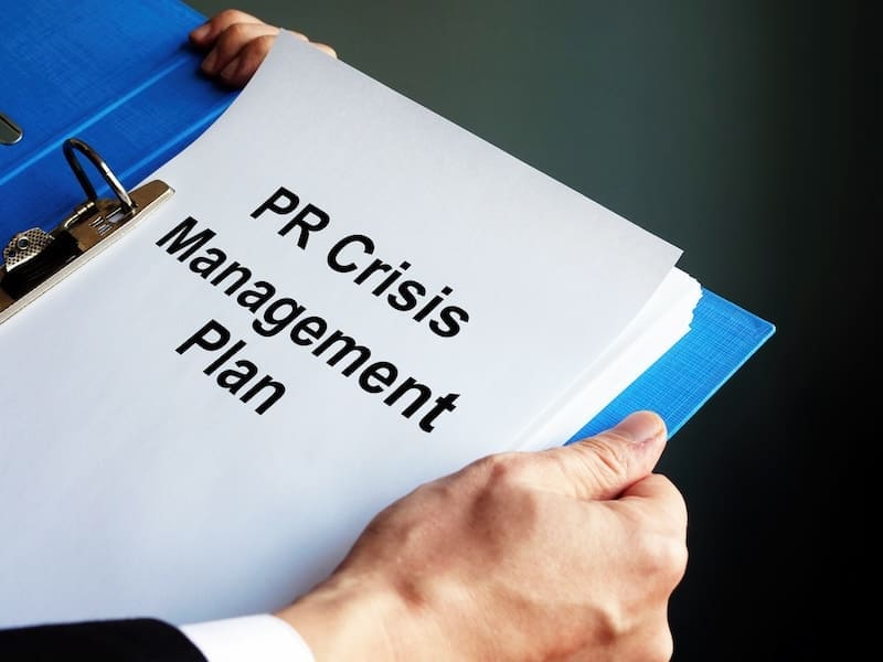 A person holding a clipboard with documents labeled "PR Crisis Management Plan." The focus is on the title of the document, suggesting the need for strategies in handling public relations crises and corporate reputation management. The individual’s hand is visible, gripping the clipboard.