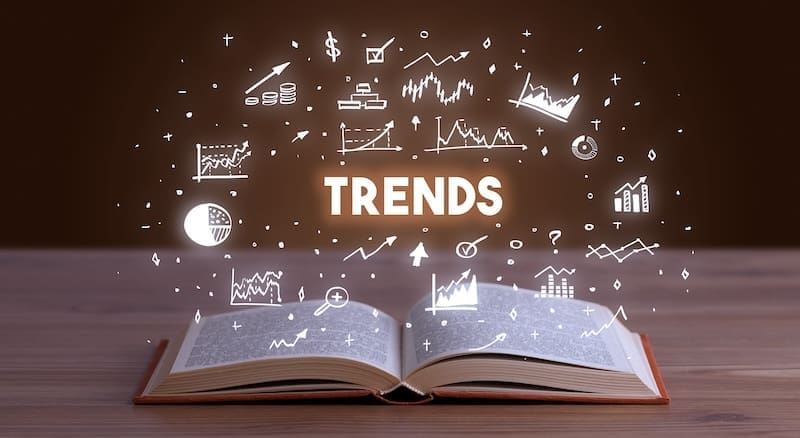 An open book lies on a wooden table with the word "TRENDS" hovering above in bold letters. Surrounding the text are various white icons depicting graphs, charts, and symbols representing business and financial trends, along with elements of corporate reputation management, all set against a blurred dark background.