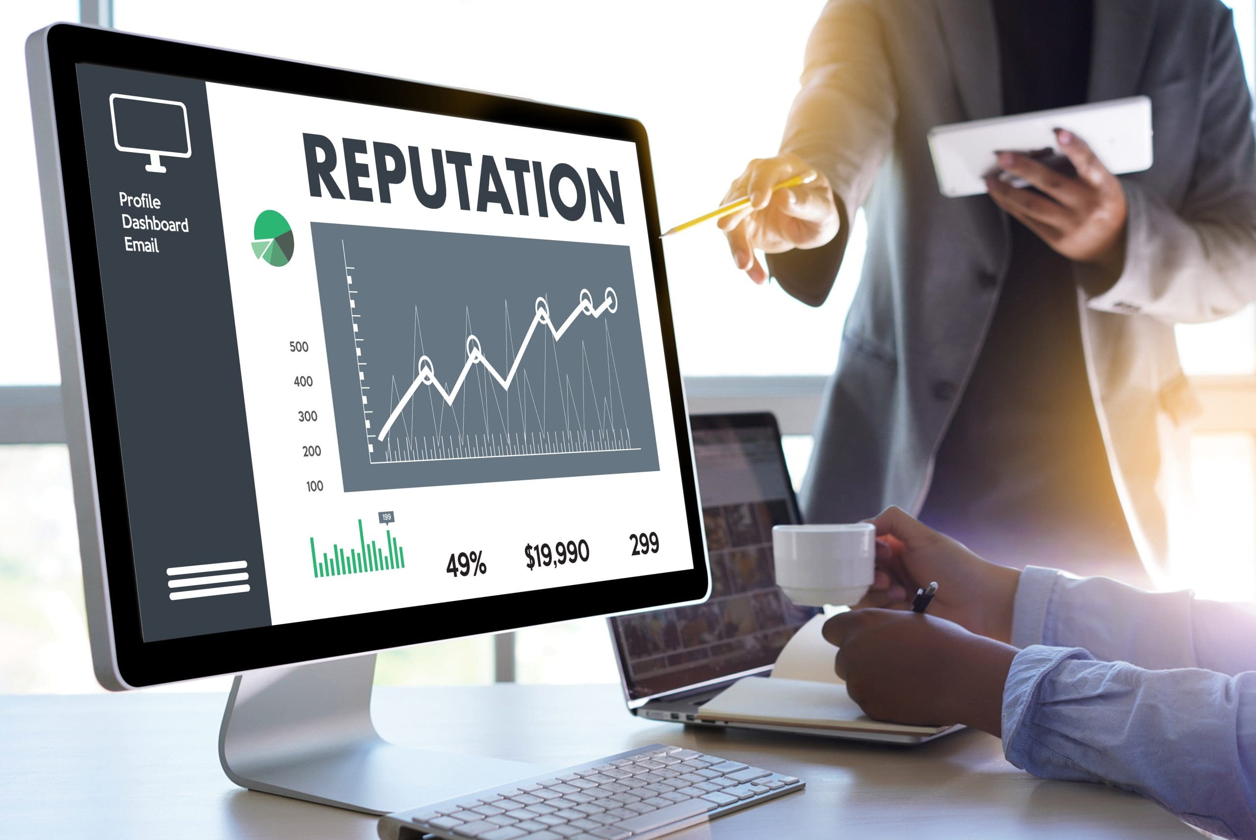 A computer screen displays a reputation analysis dashboard with graphs and statistics, essential for corporate reputation management. Two people in business attire interact; one holding a tablet points at the screen while the other takes notes and holds a cup. Sunlight streams through a window.