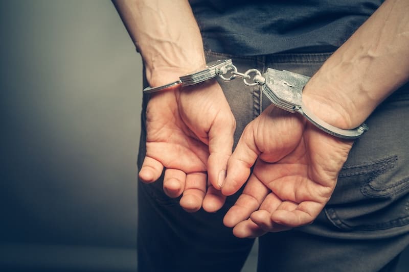 A person is shown from the back, embodying a Georgia mugshot moment, with handcuffs on their wrists. They wear a dark shirt and jeans, standing in a dimly lit environment. Their hands are positioned close together behind their back.