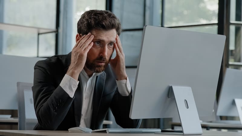 A man in a suit sits at a desk, looking stressed and holding his head with both hands in front of a computer monitor, perhaps contemplating the implications of Georgia's mugshot law. The office space around him is bright with large windows and several empty chairs.