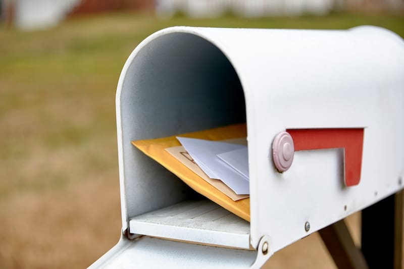 A white mailbox with the red flag lowered contains several envelopes and packages, perhaps a notice about the new Georgia mugshot law. The background is blurred, revealing a grassy area.