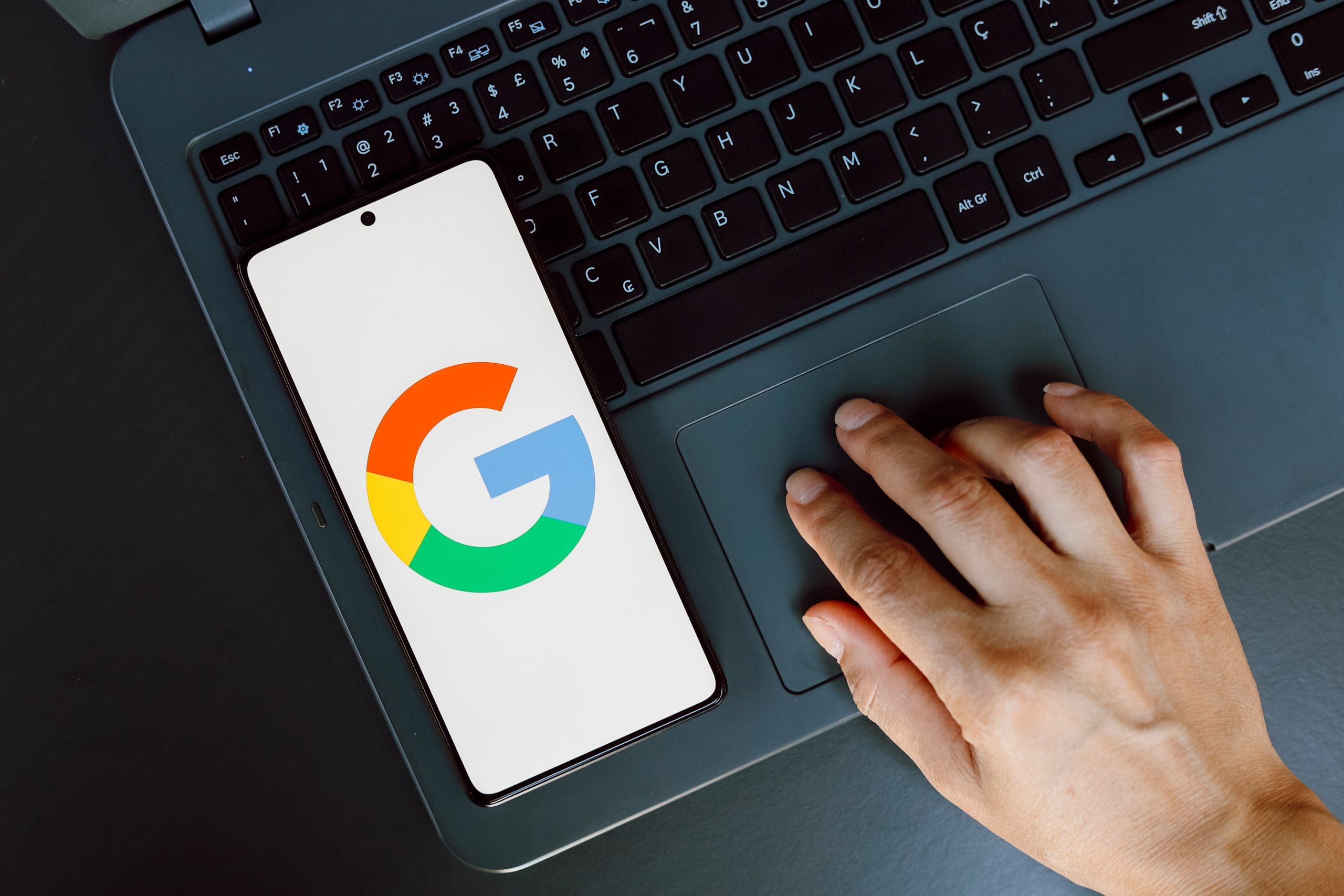 A hand uses the touchpad of a laptop while a smartphone displaying the Google logo rests on the keyboard. The laptop has a black keyboard, and the image is taken from above, focusing on the interaction between the hand, laptop, and phone.