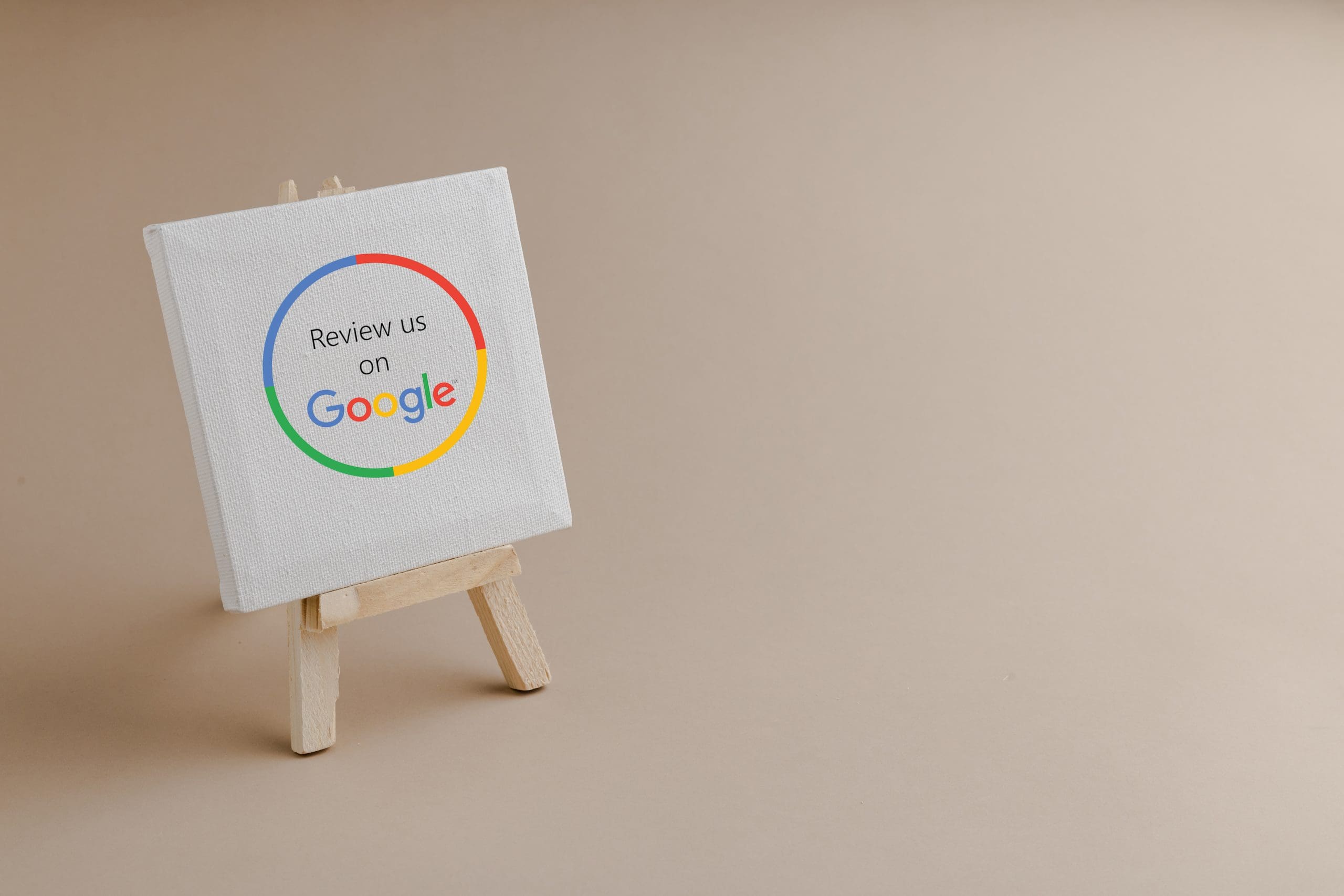 A small easel holds a white canvas with the text "Review us on Google" inside a multicolored circular design. The background is a plain beige color.