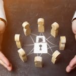 Hands encircle a group of wooden figures connected by lines, with a padlock symbol in the center. The setup suggests themes of security, protection, or network safety.