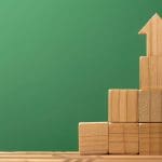 Wooden blocks are stacked in a staircase formation on a wooden surface, with an upward-pointing arrow block at the top, against a solid green background.