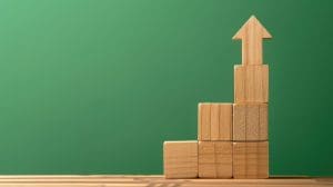 Wooden blocks are stacked in a staircase formation on a wooden surface, with an upward-pointing arrow block at the top, against a solid green background.