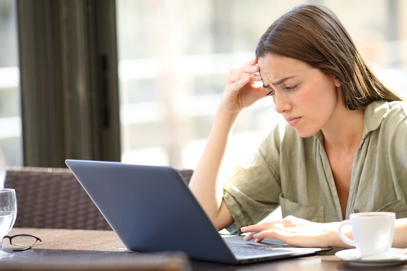 A woman with long brown hair looks concerned while researching how to get a website taken down on her laptop. Sitting indoors by a window, she wears a green shirt and sips coffee beside a glass of water.