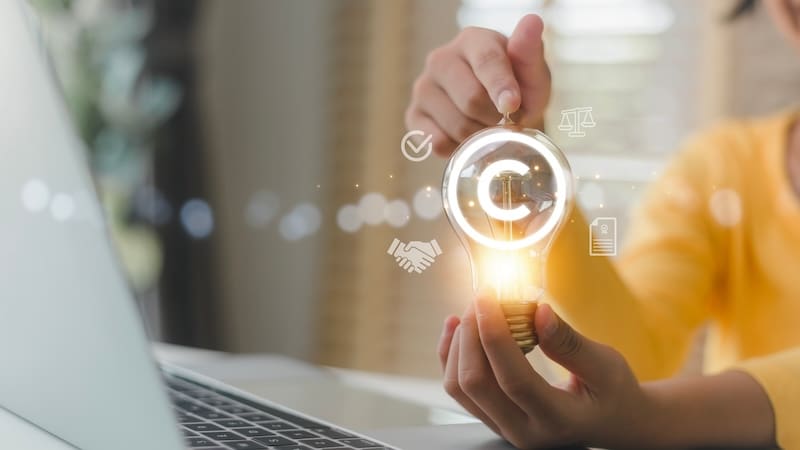 A person holds a glowing light bulb with a copyright symbol inside, surrounded by icons like a checkmark, scales, and handshake. In the foreground, a laptop hints at innovation and intellectual property concepts related to how to get a website taken down.