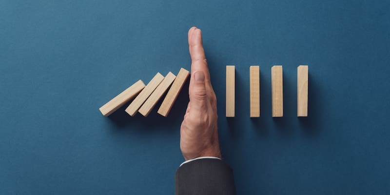 A hand in a suit jacket stops a line of wooden dominoes from falling against a blue background.