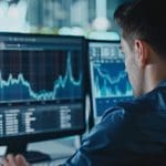 A person sits at a desk, focusing on multiple computer monitors displaying financial graphs and data charts, indicating stock market analysis. The room is dimly lit, giving an atmosphere of concentration and focus.