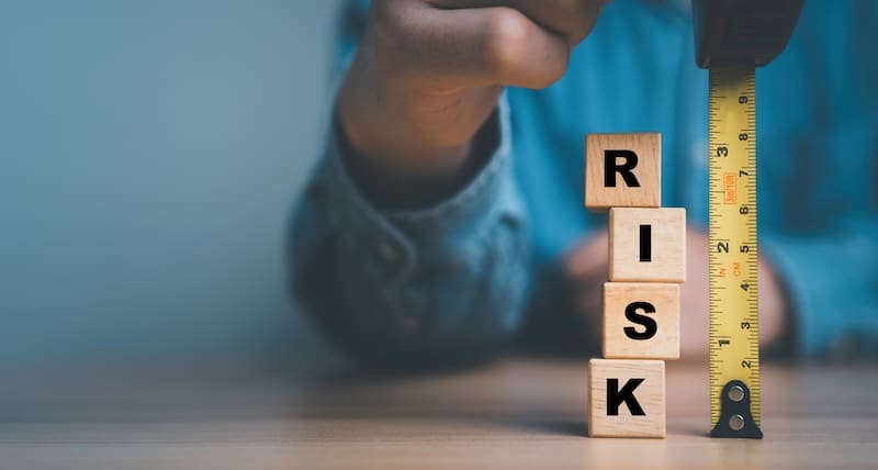 A person measures a vertical stack of wooden blocks spelling "RISK" with a yellow measuring tape. The background is blurred, and the focus is on the blocks and the measuring tape.