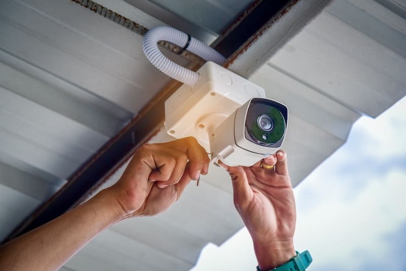 Hands of a person installing a security camera on the underside of a metal roof, with one hand holding the camera and the other securing the wires, against a background of blue sky.