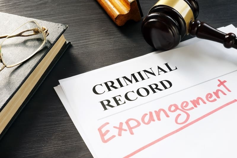 A stack of papers labeled "Criminal Record" lies on a table. The word "Expungement" is written in red across the papers. Nearby are a gavel, a pair of eyeglasses, and a stack of books.