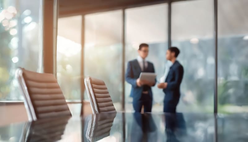 Two individuals in suits stand in an office with large windows, engaged in conversation. One holds a laptop. Foreground features blurred empty chairs and a reflective conference table. Sunlight streams in, creating a modern and professional atmosphere.