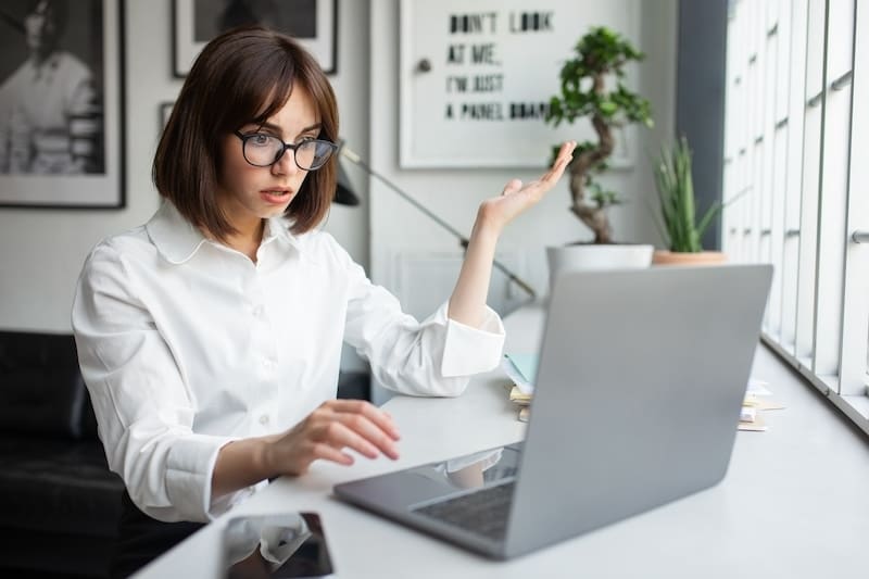 A woman with glasses, wearing a white shirt, sits at a desk looking frustrated at her laptop. Raising one hand as if questioning predictive search results, she glances around the bright room with large windows and framed pictures lining the wall behind her.