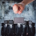 A large hand controls puppets with TV screens for heads, displaying static. The figures, dressed in black suits, stand against a grungy concrete background. The imagery suggests themes of control and media influence.