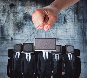 A large hand controls puppets with TV screens for heads, displaying static. The figures, dressed in black suits, stand against a grungy concrete background. The imagery suggests themes of control and media influence.