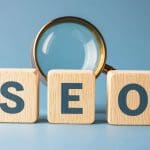 Wooden blocks spelling "SEO" sit in front of a magnifying glass, symbolizing the clarity expert SEO services bring to your digital strategy, all set against a blue background.