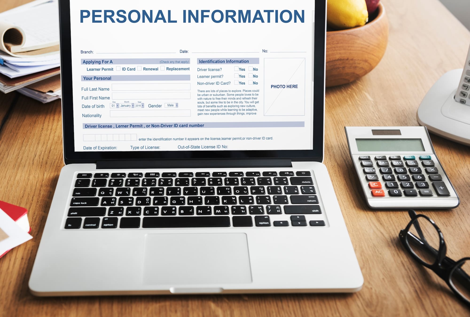 A laptop displaying a form titled "Personal Information" on the screen. The form includes fields for name, ID, and driver's license details. A calculator and a pen are on the desk beside the laptop, with papers partially visible to the left.