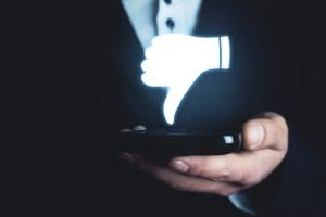 A person in a suit holds a smartphone with a glowing thumbs-down icon hovering above it against a dark background.