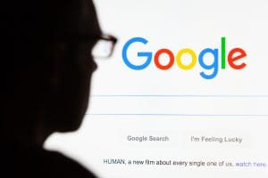 Silhouette of a person in glasses facing a computer screen displaying the Google homepage. The screen features a search bar with options "Google Search" and "I'm Feeling Lucky". Text at the bottom mentions a film titled "HUMAN".