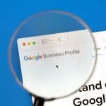 A magnifying glass focusing on the "Google Business Profile" webpage on a computer screen. The website's header is visible, displaying colorful text on a clean, white background.