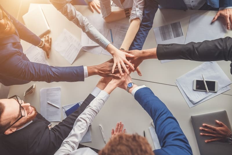 A group of diverse professionals in business attire, led by the CEO, gather around a table, stacking their hands in a gesture of teamwork and unity. Amid papers and technology, they emphasize information privacy, creating a collaborative yet secure work environment.