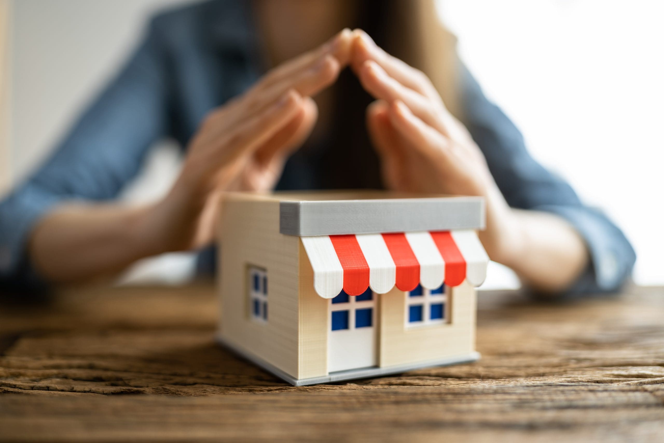 A pair of hands with fingers interlaced hover protectively over a small wooden model of a house with a red-and-white striped awning, placed on a wooden table. The person is wearing a denim shirt.