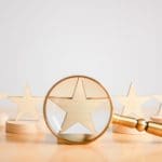 A gold magnifying glass focuses on a wooden star in the foreground, symbolizing how to deal with bad reviews. In the background, four more wooden stars are displayed on round bases, creating a row. All elements are set on a light wooden surface.