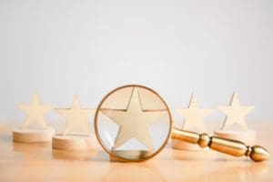 A gold magnifying glass focuses on a wooden star in the foreground, symbolizing how to deal with bad reviews. In the background, four more wooden stars are displayed on round bases, creating a row. All elements are set on a light wooden surface.