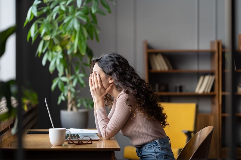 A woman with curly hair sits at a desk, holding her head in her hands in frustration. She faces a laptop, with an open notebook and a coffee mug nearby. Bookshelves and a large plant are in the background.