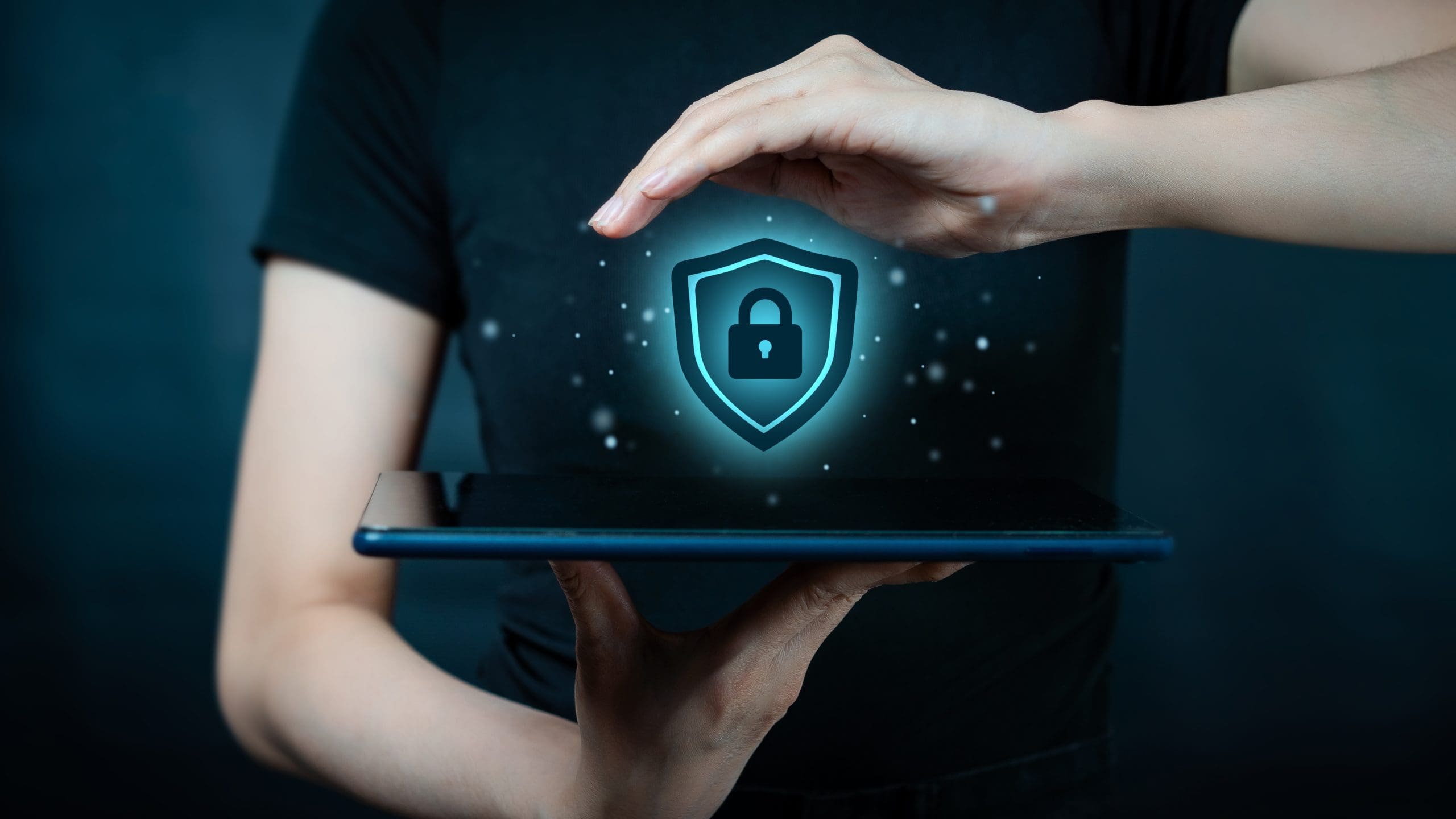 Person holding a digital tablet, with a holographic shield and padlock icon hovering above, symbolizing data security. Hands are positioned above and below the icon, creating a protective gesture. The background is dark and blurred.