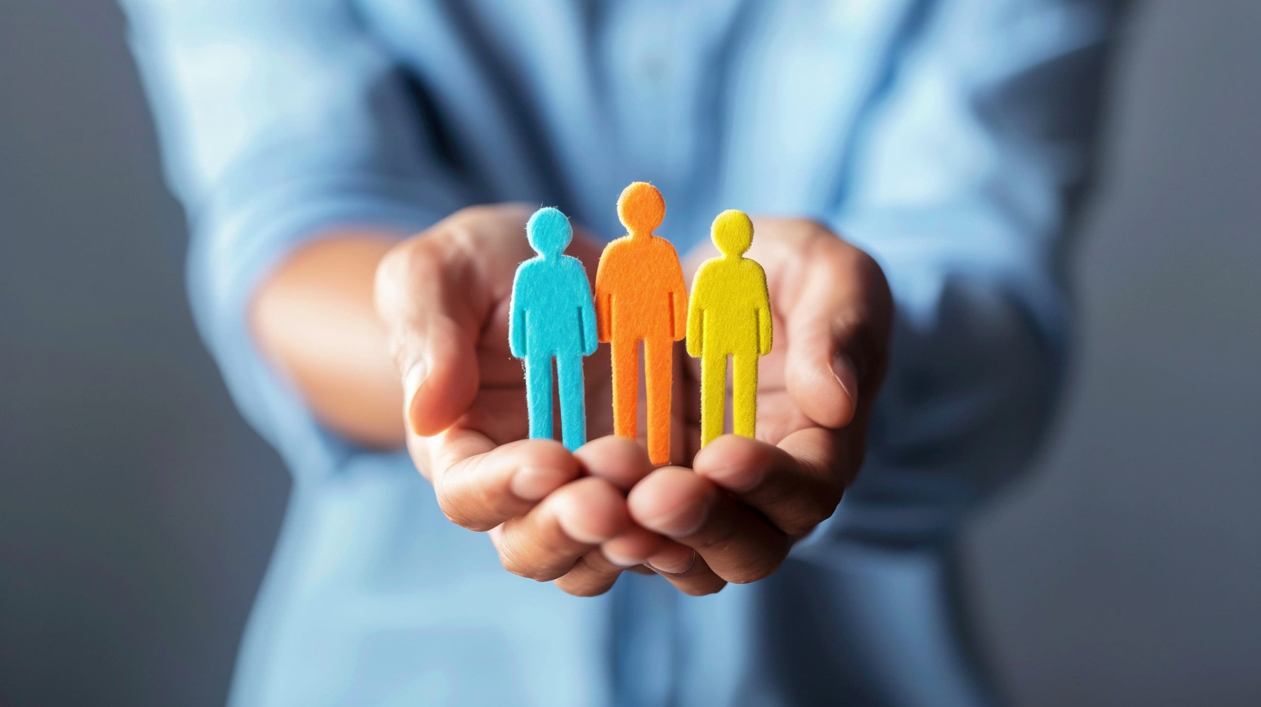 Hands hold three cutout figures in blue, orange, and yellow. The figures represent people and are symbolically positioned in the person's palms, suggesting care or protection. The background is blurred, focusing attention on the hands and figures.