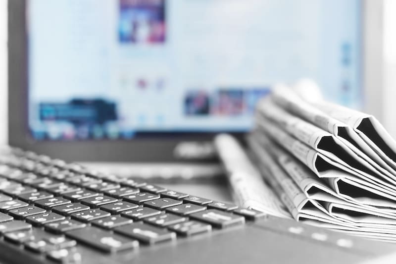 A close-up of a keyboard in front of a blurred computer screen, with a stack of folded newspapers on the right. The grayscale image subtly reflects the interplay between digital platforms and print media, echoing themes of CEO reputation management amidst evolving communication landscapes.