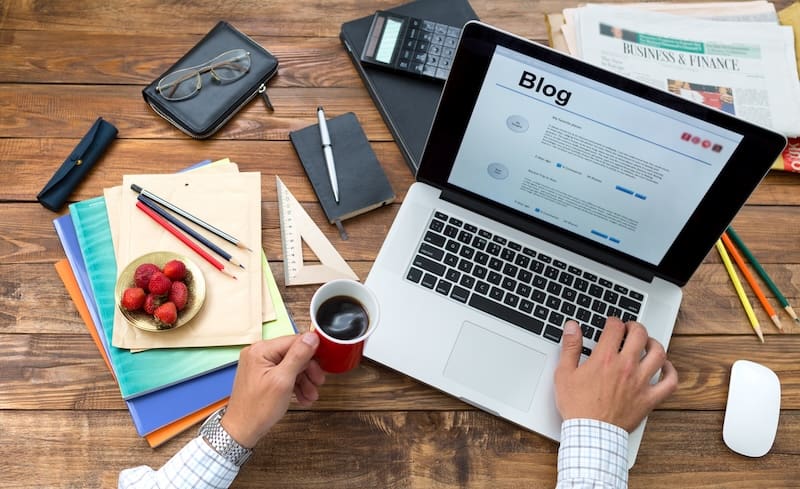 A person's hand cradles a coffee cup, poised over a laptop displaying tips to start a blog. The desk is alive with organized chaos—strewn with notebooks, papers, folders, strawberries in a bowl, a calculator, pens, and the "Business & Finance" section of the newspaper.