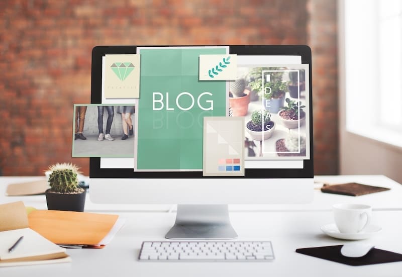 A computer monitor showcases a vibrant blog webpage with images and text, including plants, a group of people, and the word "BLOG." Enthusiastic individuals ready to start a blog will find inspiration in this setup, with a cactus, notebooks, and keyboard on the desk against a blurred brick wall backdrop.