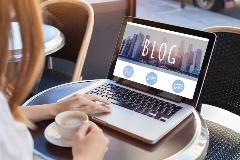 A person sits at a round table holding a cup of coffee while using a laptop to start a blog. The screen displays the word "BLOG" against a city skyline background, accompanied by three icons: an envelope, an open book, and a bar chart.